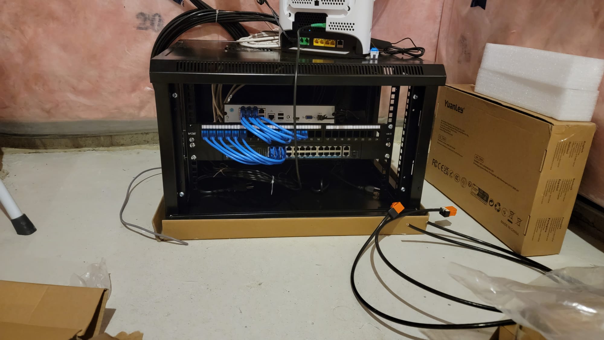 Open wall-mount network cabinet with patch panel, PoE switch, and blue Cat6 patch cables next to Yuanley equipment box during basement rough-in