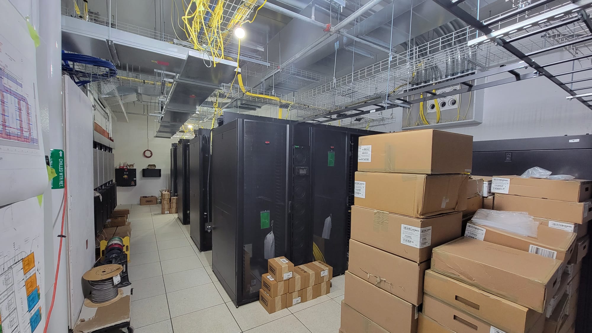 Commercial data center room with enterprise server cabinets, overhead yellow fiber tray system, and equipment crates during active build-out