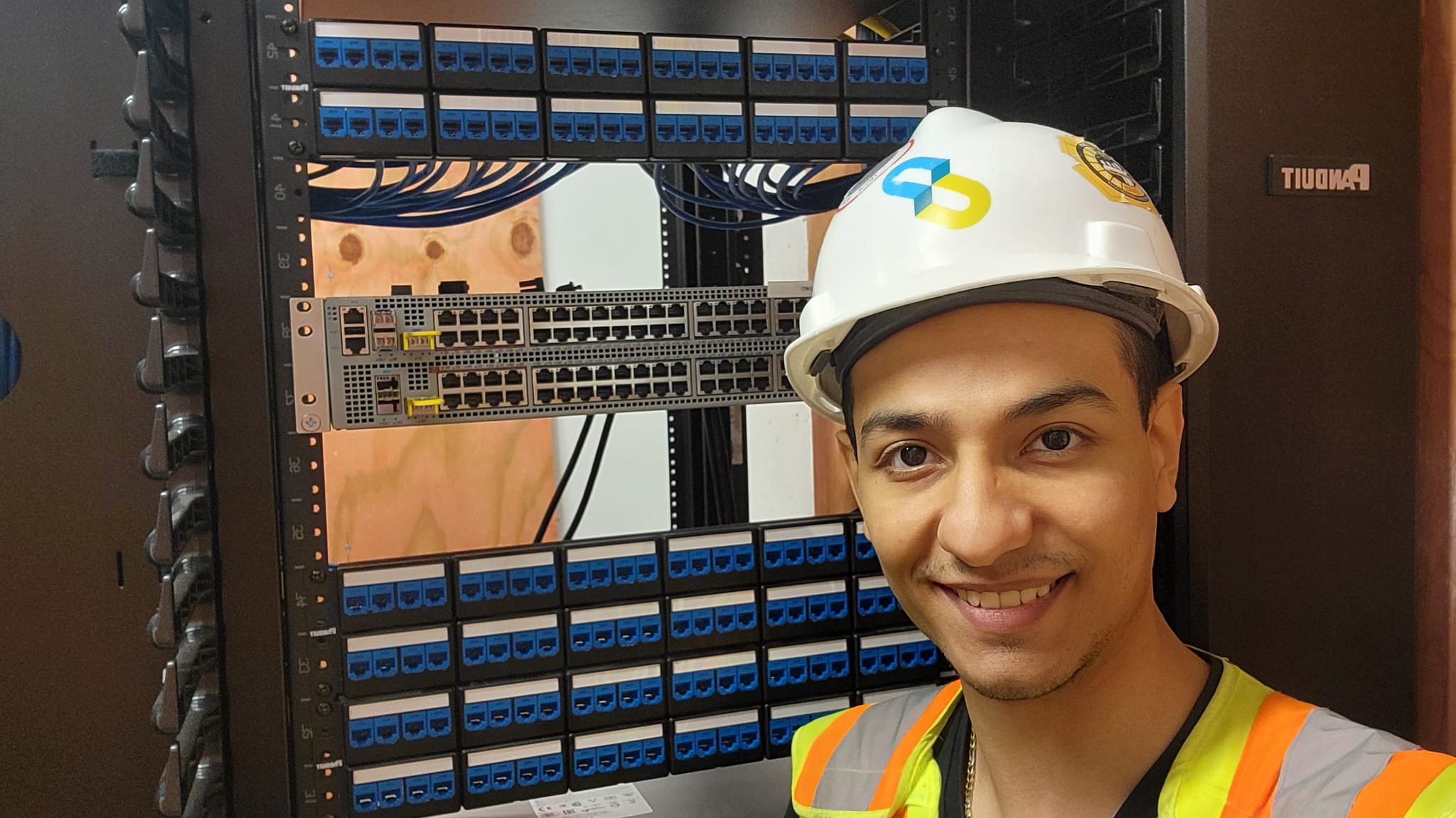 Field portrait of Ibram in branded planGroup hardhat in front of structured cabling rack with stacked switches and blue patch panels